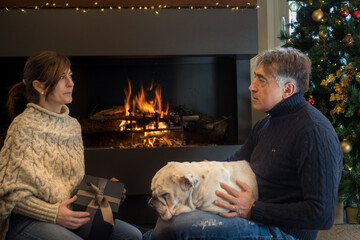 Middle-aged couple exchanges Christmas presents in front of the fireplace with their dog in the living room of the house.