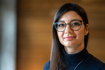 Front view portrait of young adult caucasian happy joyful woman with black long hair and eyeglasses looking to the camera while standing indoor in day with copy space real people