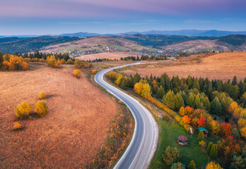 Fototapeta premium Aerial view of mountain road in forest at sunset in autumn. Top view from drone of road in woods at dusk. Beautiful landscape with roadway in hills, trees, meadows, fields, violet sky in fall. Travel