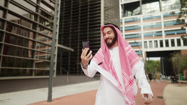 Traditional dressed happy arab man talks with family online via smartphone while walking city street