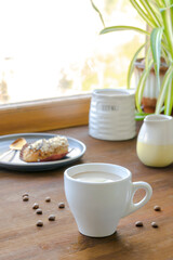 white espresso coffee cup, fresh baked eclair sweet dessert on plate kitchen table against window,...