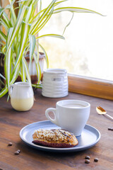 white espresso coffee cup, fresh baked eclair sweet dessert on plate kitchen table against window, utensils dishware, home green plant. morning french home breakfast