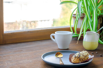 white espresso coffee cup, fresh baked eclair sweet dessert on plate kitchen table against window, utensils dishware, milk jug, home green plant. morning french home breakfast, copy space