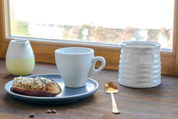 white espresso coffee cup, fresh baked eclair sweet dessert on plate kitchen table against window, utensils dishware, milk jug, home green plant. morning french home breakfast, copy space