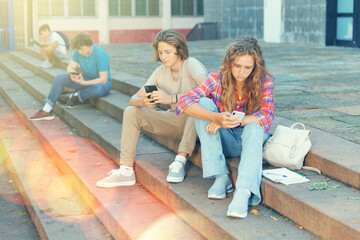 Teenage schoolers, boy and girl, using their gadgets outside school on sunny autumn day