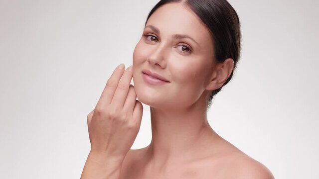 Beauty Caucasian face. Woman touching her face skin. Beautiful smiling female with fresh glowing hydrated facial skin and natural makeup on light background at studio. Skin care concept.