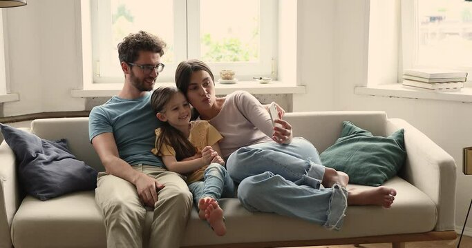 Friendly young mom dad small girl cuddle rest on comfy couch engaged in shopping online on cell watch photo video at social media. Happy family parents little child have fun with phone at home use app