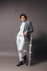 Tall handsome man dressed in white turtleneck, white jeans and grey jacket posing on the grey...