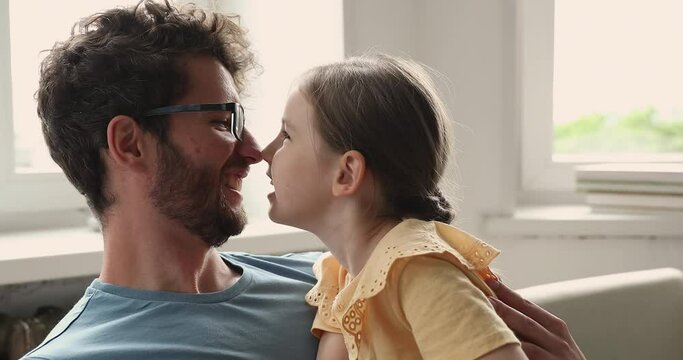 Affectionate little girl kid cuddling with beloved daddy on comfy couch playing cute game messing touching noses. Happy carefree young single father and preschool daughter kidding having fun on sofa