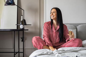 Happy young domestic woman in red pajamas drinking champagne wine glass sitting in comfortable bed