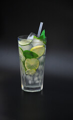 Tall glass of fresh Mojito with ice, leaves, mint, lime slices and a straw on a black background.