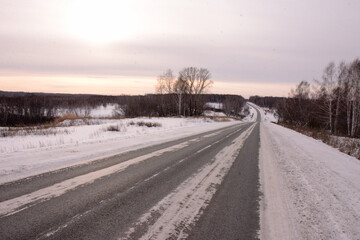 Fototapeta na wymiar A turn on an asphalt road with snowdrifts along the sides stretches across a snow-covered field to meet the setting sun.