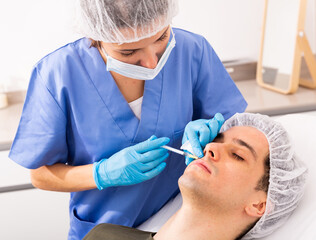 Professional female beautician performing face contouring injections to male patient at cosmetology...