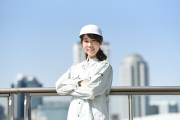 空とビルを背景に佇む作業服を着た腕組みをする若い女性