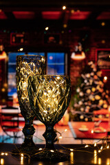 Two wine glasses, of different heights, made of curly blue glass, filled with shining lights. Shining glasses with bright lights create a festive atmosphere and a desire to celebrate Christmas and New