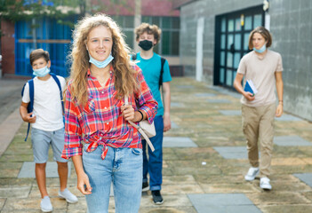Teenager girl in face mask going home after classes in school with other teens who walking in background. .