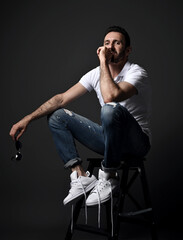 Thoughtful adult unshaved man in white polo shirt, modern torn jeans and sneakers sits on high chair, stepladder looking at camera over dark background. Side view