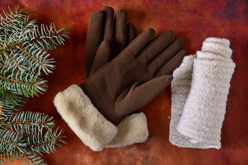 Winter woman clothing accessories. Brown soft leather gloves with fur trim and rolled white down knitted warm scarf next green twig of fir christmas tree on brown background. Flat lay