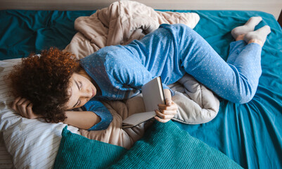 Caucasian woman reading book in the morning at home in the living room, lying in bed in sleeping clothes. Bedtime. Curly hair