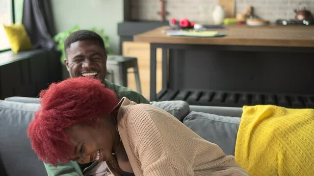 Young woman, man having online talk or taking selfie in front of phone screen on couch at home spbd. Close-up view of American african couple holds smartphone in hand and poses for camera, laughs and