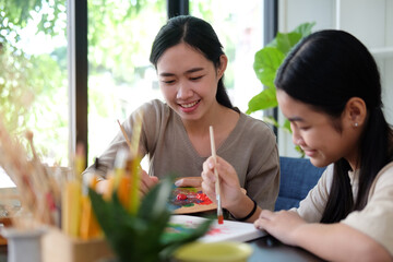 Happy young Asian mother and her daughter painting together at home.