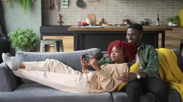 Young couple in love using smartphone and lying on couch in apartment room spbd. American African woman, man hold phone in hand and look at screen with smiles, guy hugs female and sits on sofa in