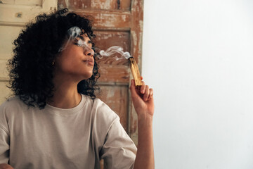 Young mixed race woman holding a smoking palo santo smudging herself and the room. Copy space.