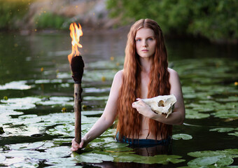 Beautiful red haired woman with animal skull and flaming torch in hands standing in water with lily...