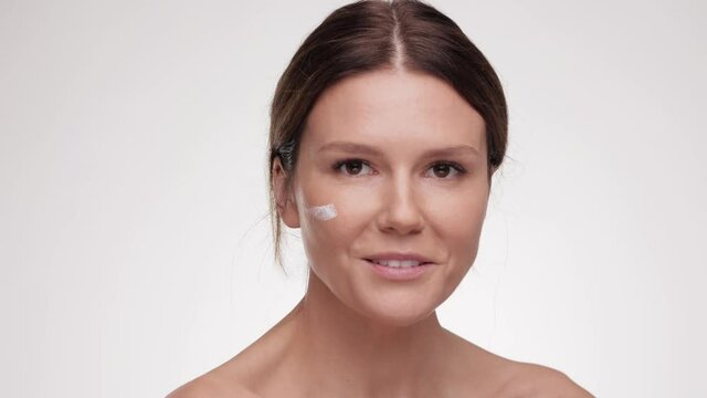 Female beauty procedures. Lady cream applying smear of pampering cream on her cheek and smiling to the camera, white studio background. Skin care concept. Cosmetic Concept.