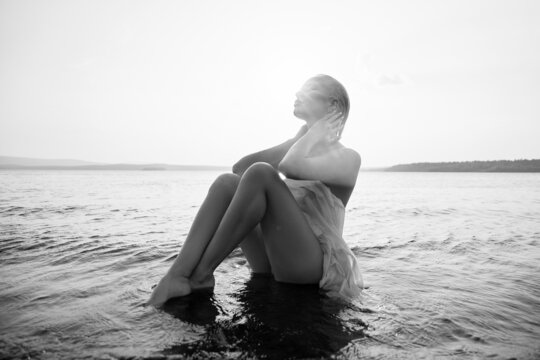 Art nude sexy blonde with short haircut is sitting in water on shore beach of lake at sunset. Wet hair and a woman body. Secluded beach holiday. Black and white - Powered by Adobe