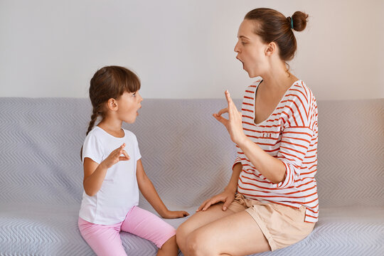 Indoor shot of female physiotherapist posing with small child girl indoors while sitting on sofa, fix speech problems, demonstrates how to pronounce sounds.