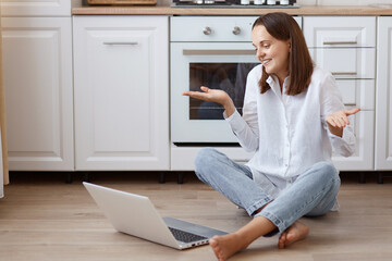 Uncertain dark haired Caucasian female sitting on floor in kitchen, looking at laptop display with confused facial expression and spreading hands aside, wearing white shirt and jeans.