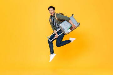 Portrait of jumping young excited Asian tourist man with baggage in isolated studio yellow...