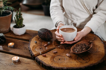 Hot handmade ceremonial cacao in white cup. Woman hands holding craft cocoa, top view on wooden...