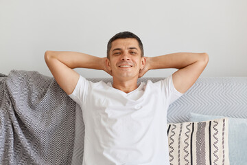 Closeup portrait of satisfied Caucasian brunette man wearing white t shirt, posing light room while sitting on gray couch, posing with raised arms, having confident facial expression.