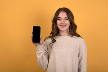 A young and happy Caucasian brunette girl with wavy hair in a casual jumper is holding a smartphone and showing a display with copy space on an orange studio background.