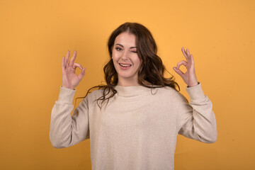 Happy and attractive caucasian brunette girl with wavy hair in a casual jumper showing okay gesture with her hands on an orange studio background.