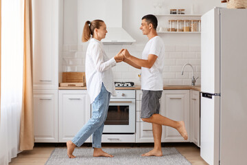 Obraz premium Portrait of happy positive young adult Caucasian man and woman, posing indoor, dancing in kitchen, celebrating their anniversary, wearing casual style attires.
