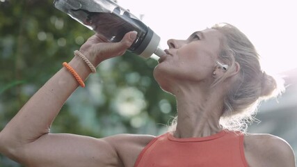 Aged woman in sport clothes drinking water after running