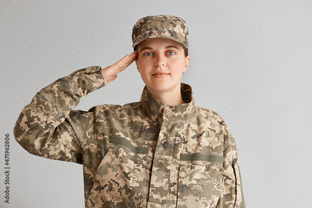 Wall mural Indoor shot of positive woman soldier wearing camouflage uniform and cap, saluting with positive facial expression, standing against light background, being glad to serve in army.