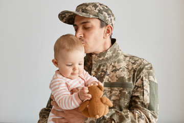 Indoor shot military man wearing camouflage uniform and cap, standing with his little infant daughter in hands, father missing his child and kissing baby, expressing love.