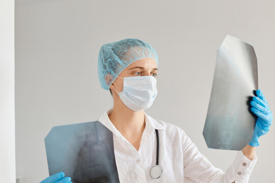 Indoor shot of female doctor looking at an x-ray, examining xray picture, woman wearing medical cap, surgical mask and gown working in clinic or hospital.