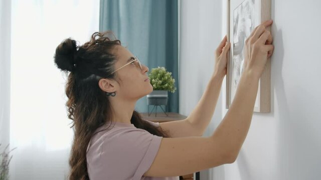 Slow motion of young creative mixed race woman homeowner decorating wall with picture and looking busy with household activities. People and lifestyle concept.