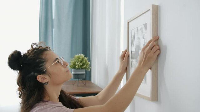 Close-up of young Asian woman interior designer hanging modern picture on wall indoors in apartment. House decoration and interior elements concept.