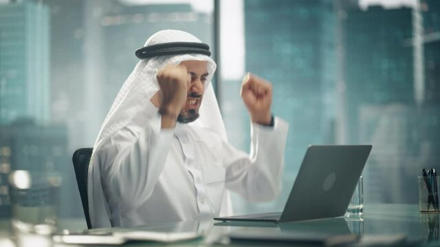 Arab Businessman in White Traditional Outfit Sitting in Office and Working on Laptop Computer. Business Manager Celebrates Successful Investment Deal. Saudi, Emirati, Arab Businessman Concept.