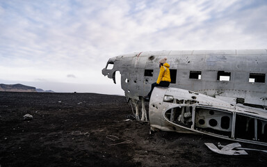 Alone tourist sitting on the wing of an airplane, wreckage from an airplane crash in Iceland on Solheimasandur black sand beach.  Abandoned DC 3 plane. Popular Travel destinations.