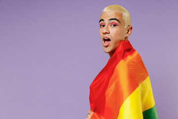 Young side view shocked latin gay man 20s with make up wrapped in rainbow striped flag look camera isolated on plain pastel purple background studio portrait. People lifestyle fashion lgbtq concept