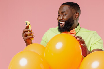 Young black gay man 20s wear green t-shirt bow tie hold bunch of air inflated helium balloons celebrating birthday party hold in hand use mobile cell phone isolated on plain pastel pink background