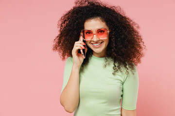 Young curly latin woman 20s years old wears mint t-shirt sunglasses hold use talk on mobile cell phone conducting pleasant conversation isolated on plain pastel light pink background studio portrait.