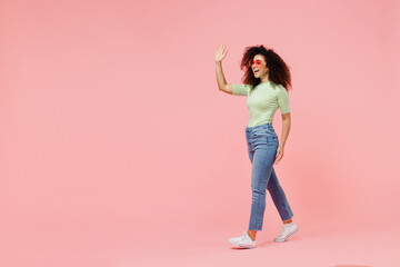 Full size body length young curly latin woman 20s wears casual clothes sunglasses go walk move meet greet waving hand as notices someone isolated on plain pastel light pink background studio portrait.